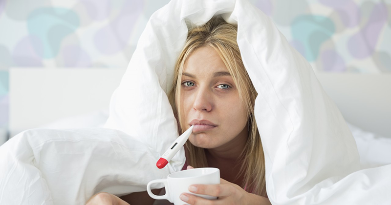 Portrait of sad woman with coffee mug taking temperature while wrapped in quilt on bed --- Image by © moodboard/Corbis