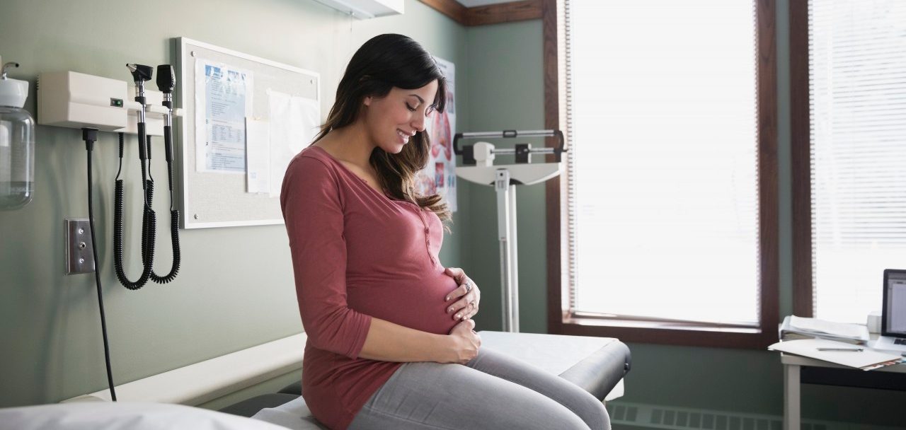 10 Dec 2014 --- Smiling pregnant woman holding stomach in examination room --- Image by © Hero Images Inc./Hero Images Inc./Corbis