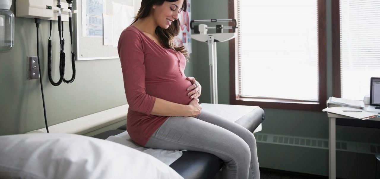 10 Dec 2014 --- Smiling pregnant woman holding stomach in examination room --- Image by © Hero Images Inc./Hero Images Inc./Corbis