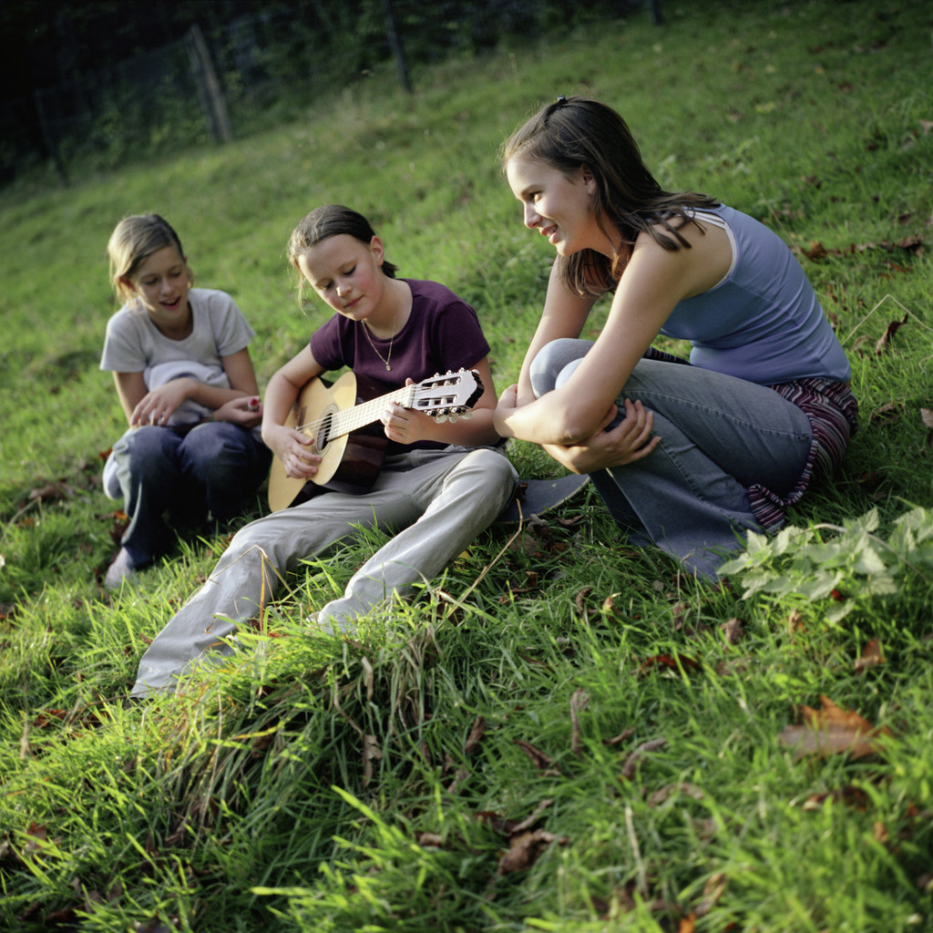 Three Girls Sitting with Guitar in Meadow --- Image by © Mika/Corbis