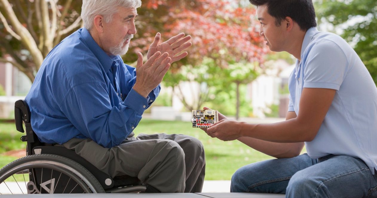 Massachusetts, USA --- Engineer with muscular dystrophy and diabetes in his wheelchair talking with design engineer about microchips on circuit board --- Image by © Mark Hunt/Huntstock/Corbis