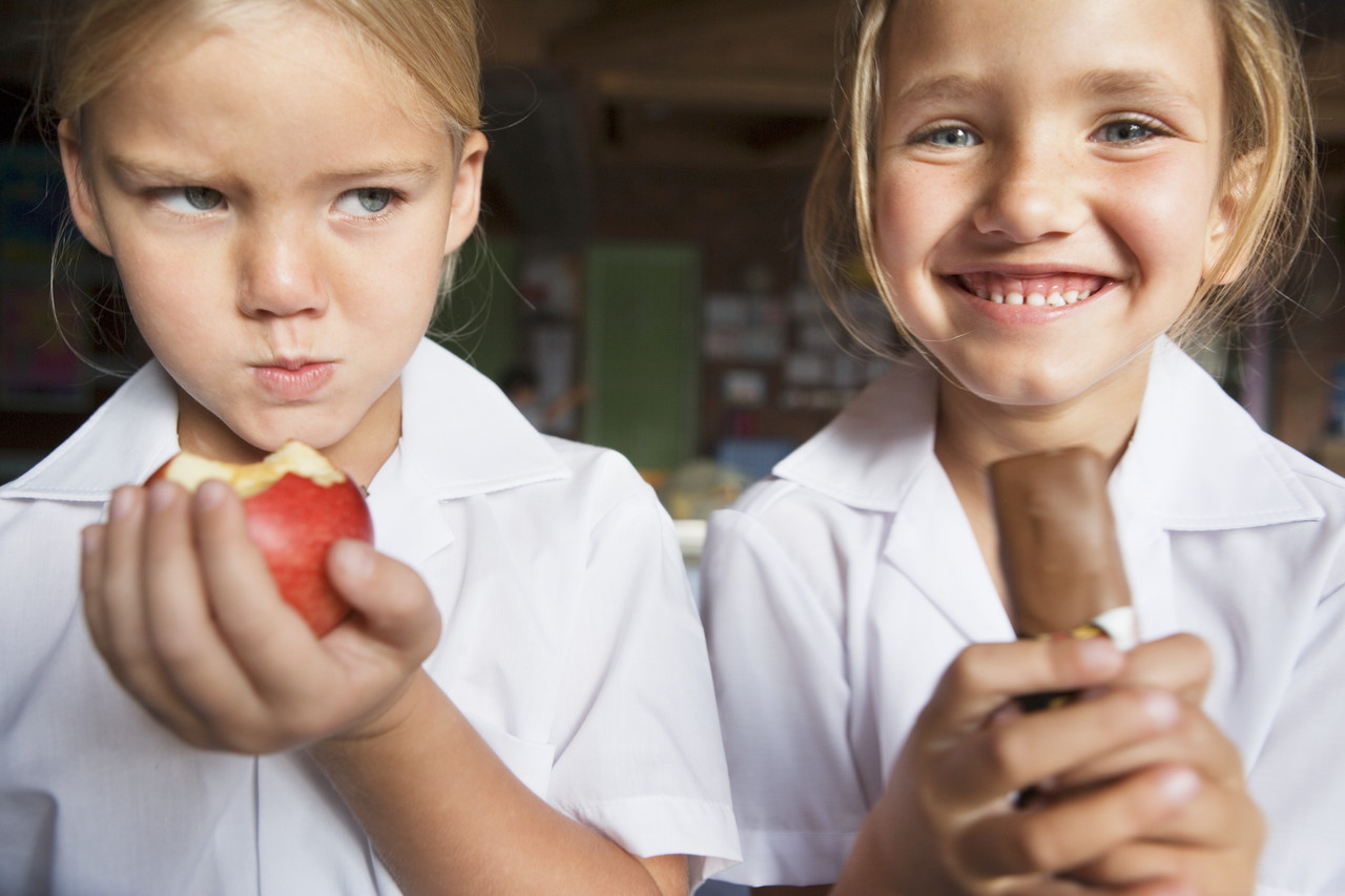 Angry Sibling Holding an Apple While Her Twin Eats Chocolate --- Image by © Roy McMahon/Corbis