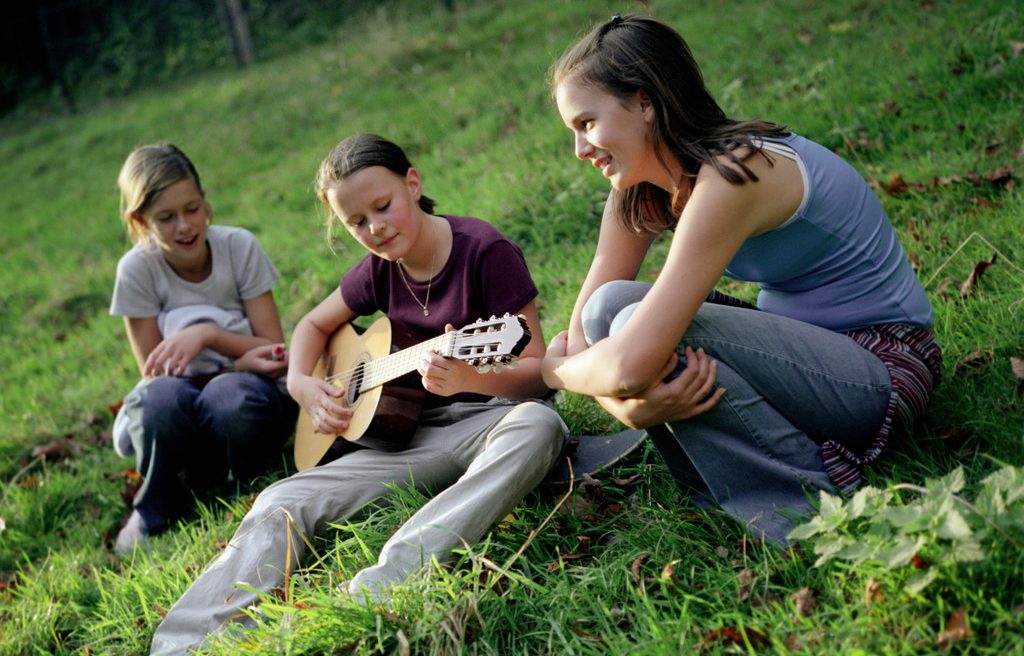 Three Girls Sitting with Guitar in Meadow --- Image by © Mika/Corbis